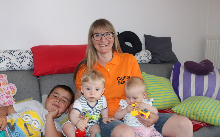 Auf dem Bild sieht man eine lachende Frau, die auf einer Couch mit 3 Kleinkindern sitzt