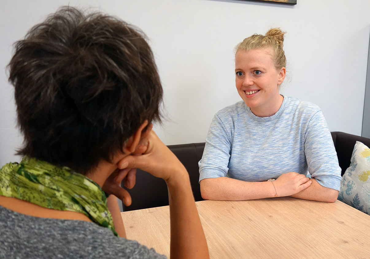 Camilla sitzt im Hintergrund, sie sieht eine Frau an, von der man nur den Rücken sieht